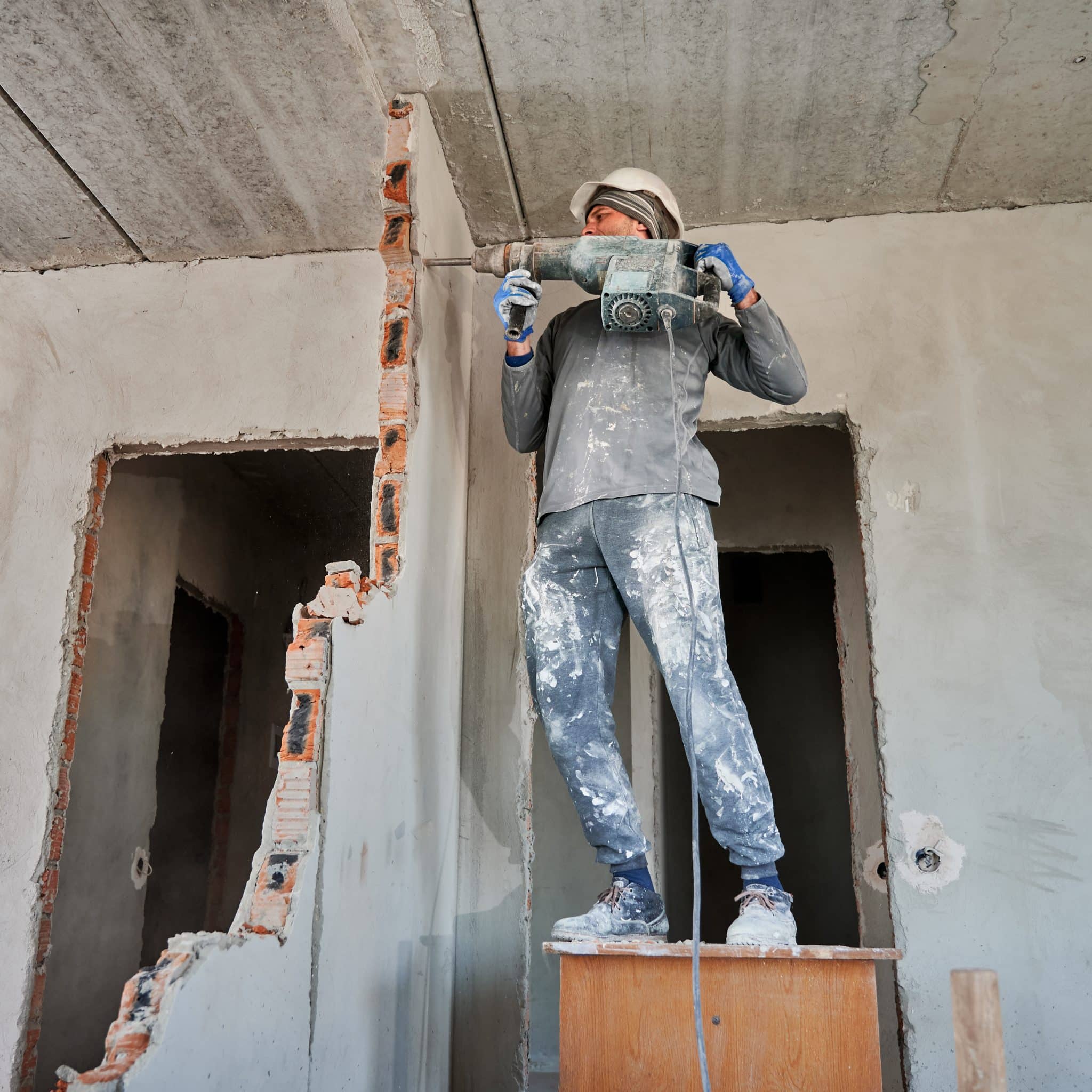Full length of man in workwear drilling wall with hammer drill. Male worker using drill breaker while destroying wall in apartment under renovation. Demolition work and home renovation concept.