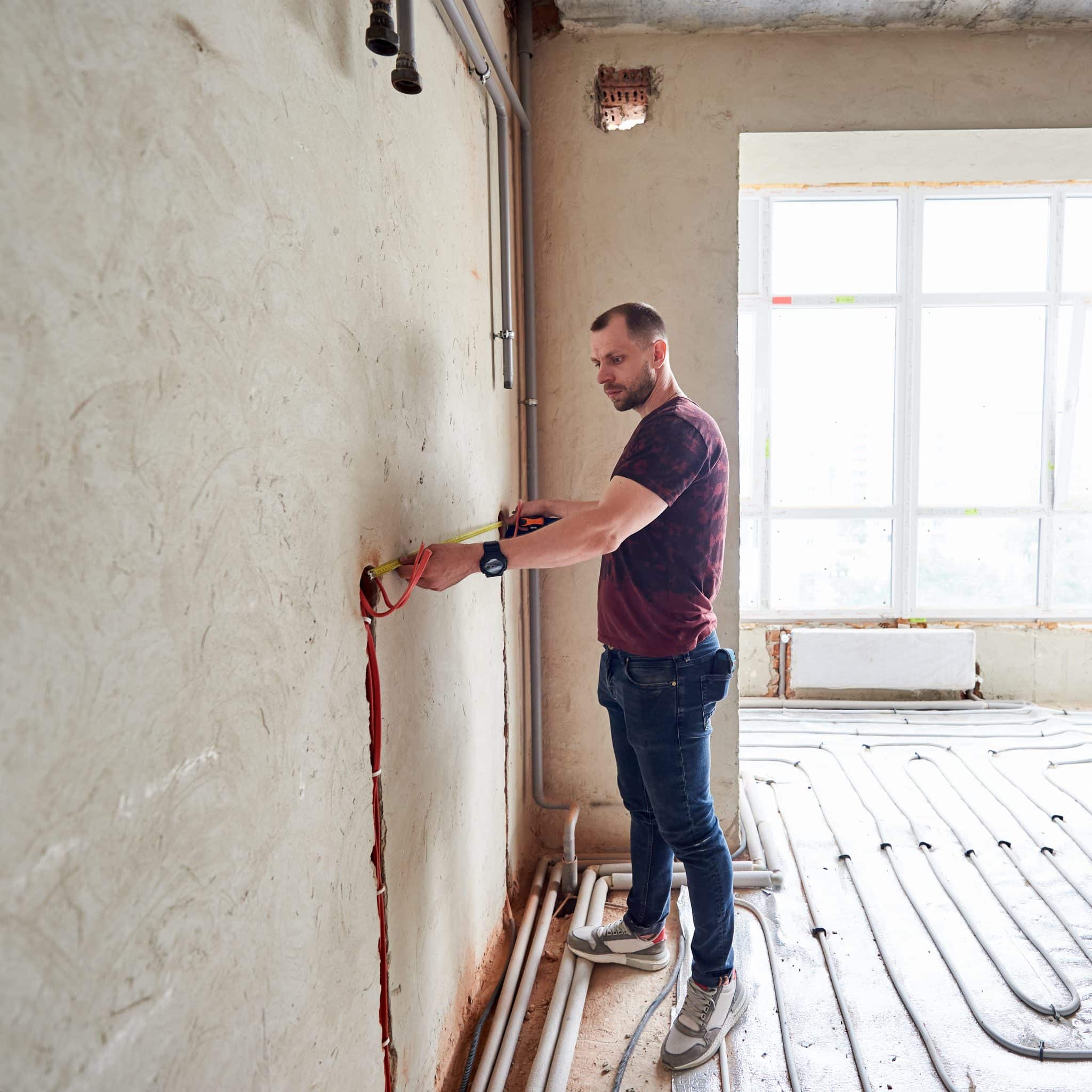 Young man in empty unfinished apartment measuring the wall with a help of measure tape, standing on floor heating pipe system against panoramic window. Concept of refurbishment and renovation.